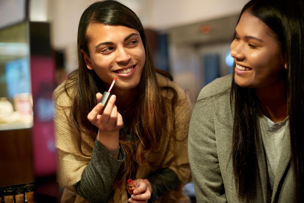 Smiling gender fluid person trying on lipstick with their friend in a cosmetics shop.