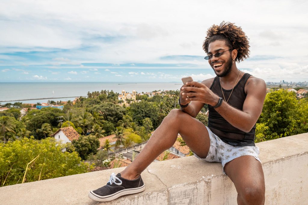 A young man in shorts smiling while sitting on an outdoor wall and holding his mobile phone with the beach and ocean behind him.