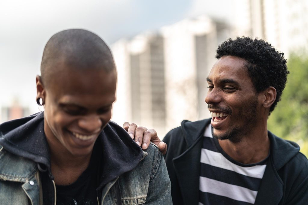 Close-up photo of two young men outdoors in an urban location laughing while one places his hand on the other's shoulder.